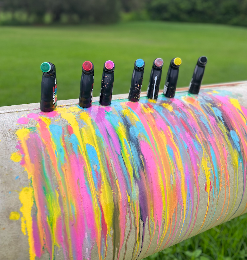 POSCA MOP'R markers lined up above multicoloured drip artwork on outdoor concrete